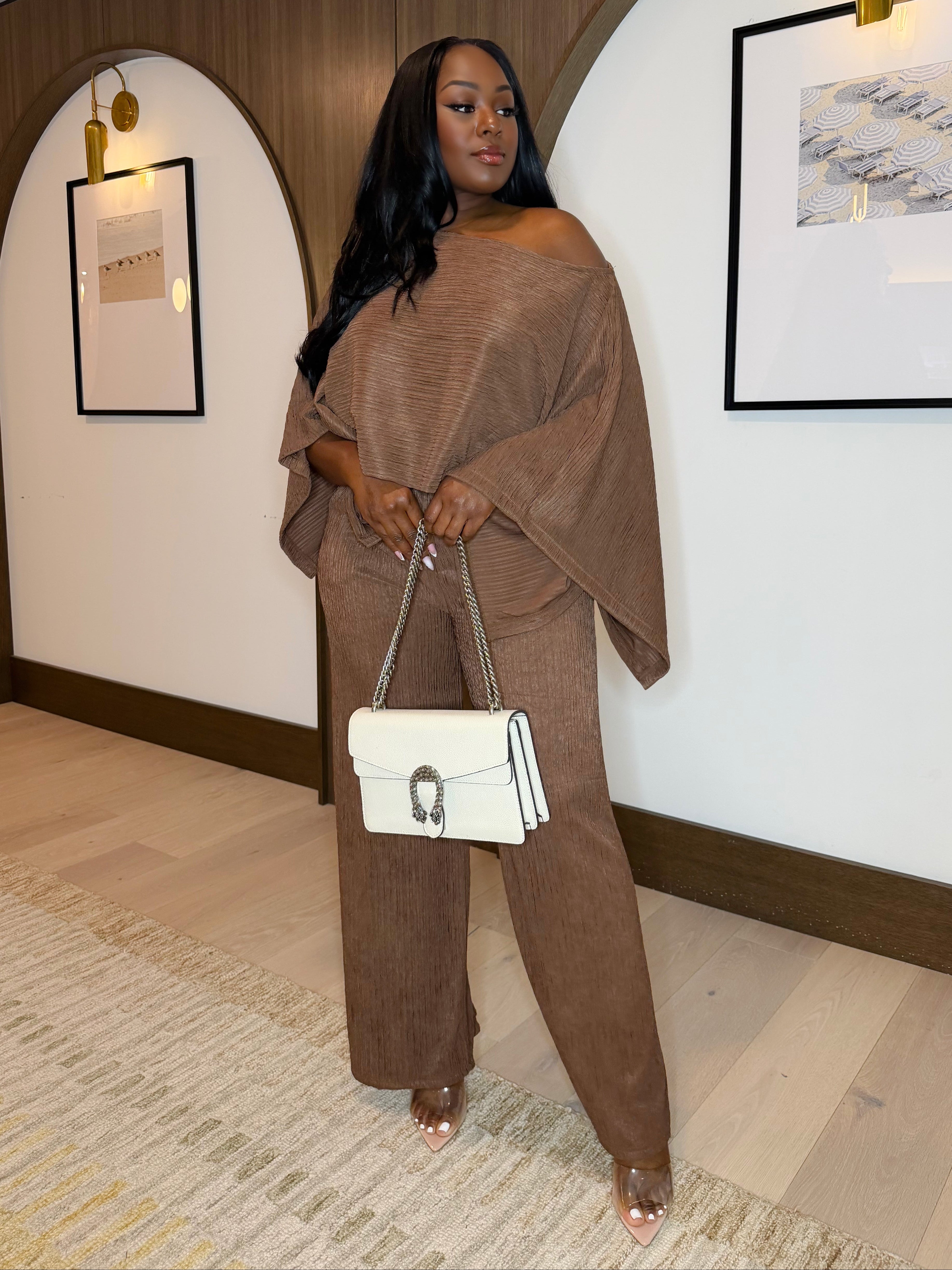 Woman in a brown outfit holding a white handbag in an indoor setting with framed pictures on the wall.
