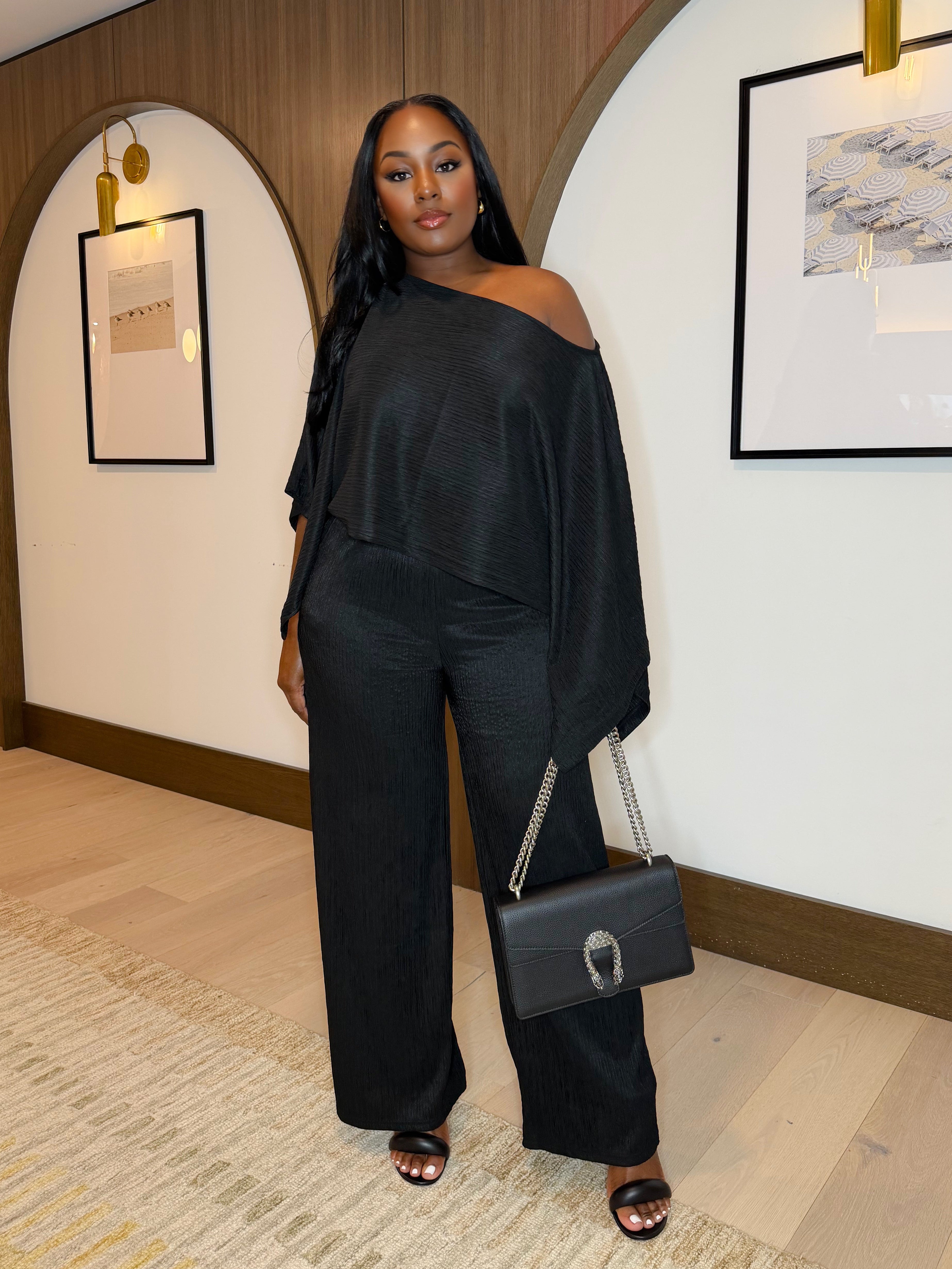 Woman in a black outfit holding a handbag in an indoor setting with framed pictures on the wall.