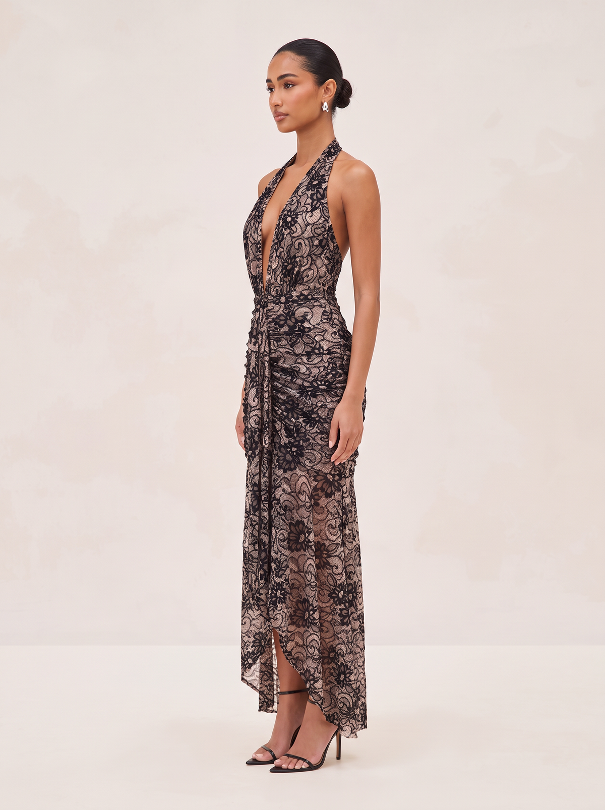 A woman modeling a sleeveless halter-neck maxi dress with a deep plunging neckline, featuring a black and brown floral lace overlay over a sheer nude underlayer, set against a warm beige studio background.