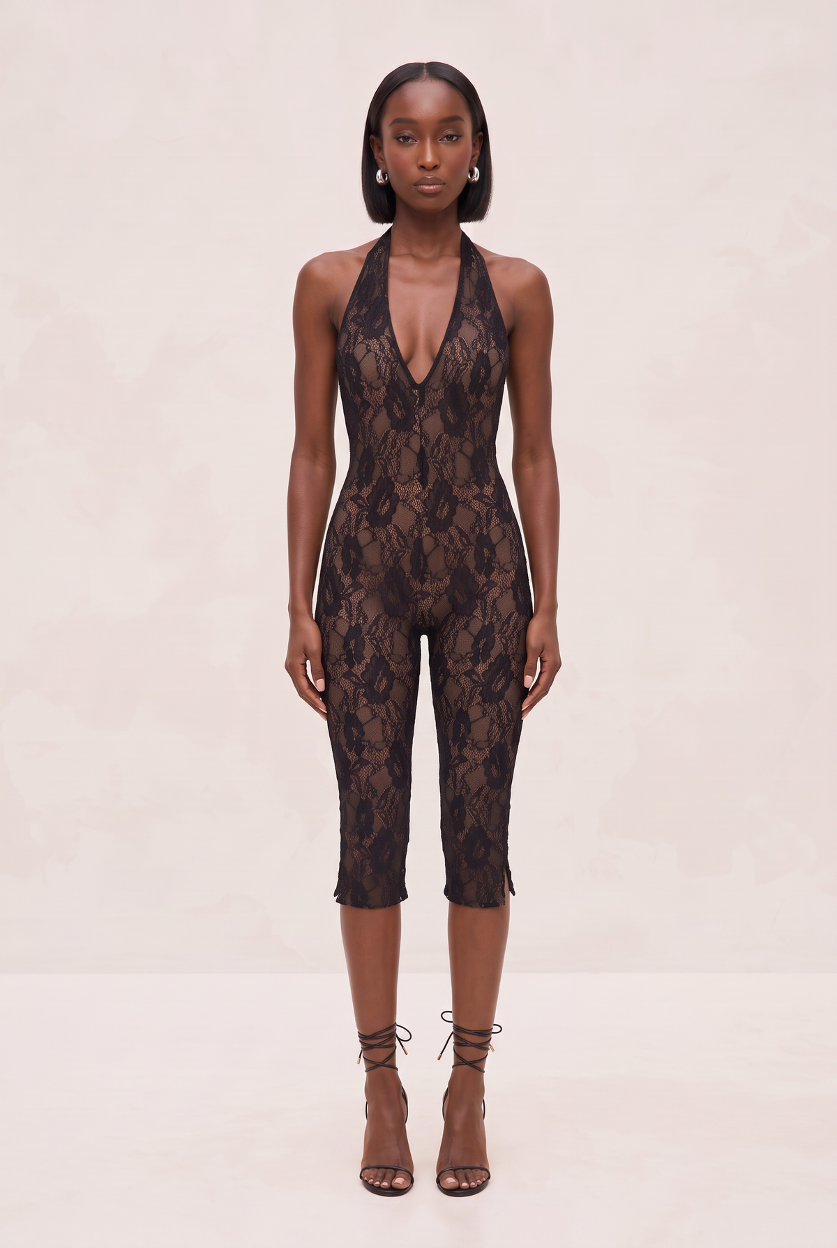 A model stands facing forward wearing a black lace halter-neck jumpsuit with a deep plunging neckline and capri-length legs, paired with brown lace-up heeled sandals against a plain beige studio backdrop.