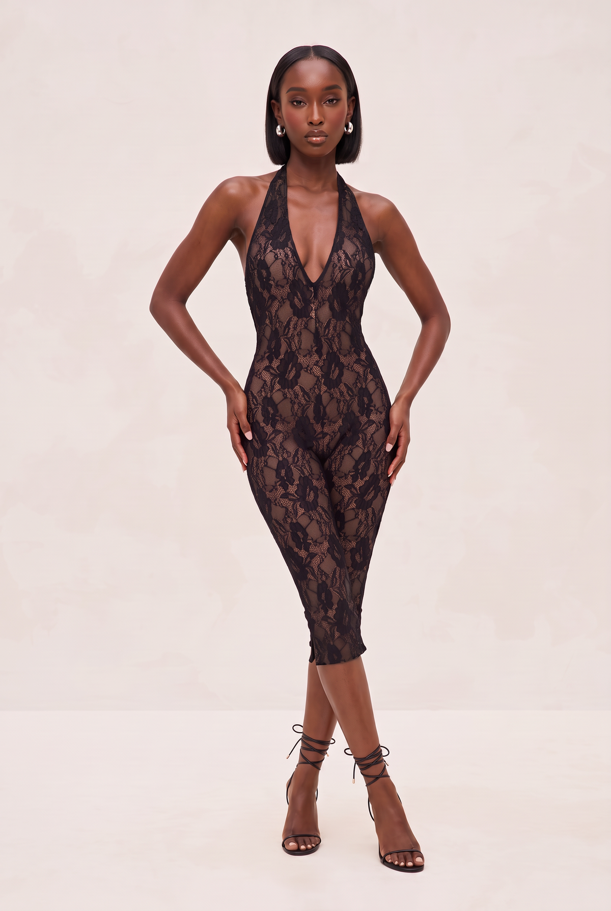 A model wearing a black lace halter-neck jumpsuit with a plunging neckline and mid-thigh length, paired with black lace-up high-heeled sandals, set against a plain light beige studio background.