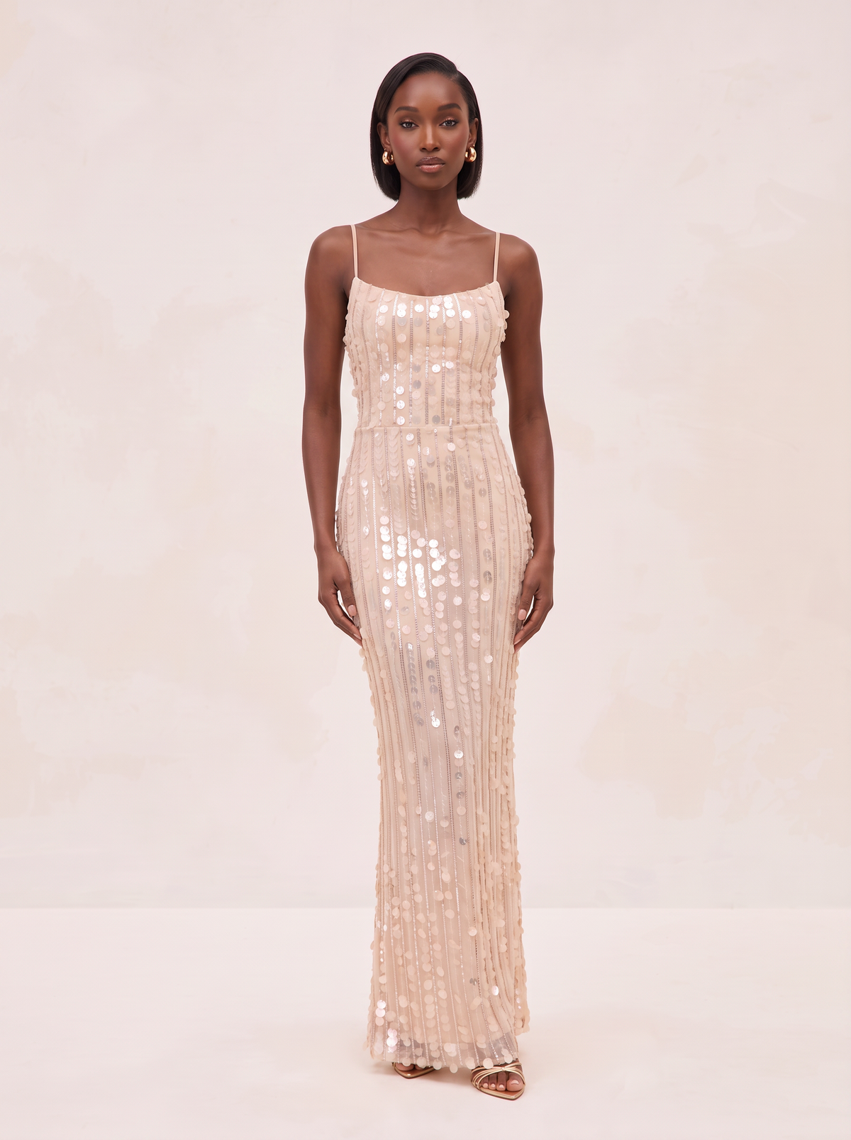 A woman modeling a floor-length champagne sequined gown with thin spaghetti straps and a fitted silhouette, standing against a warm beige studio backdrop.