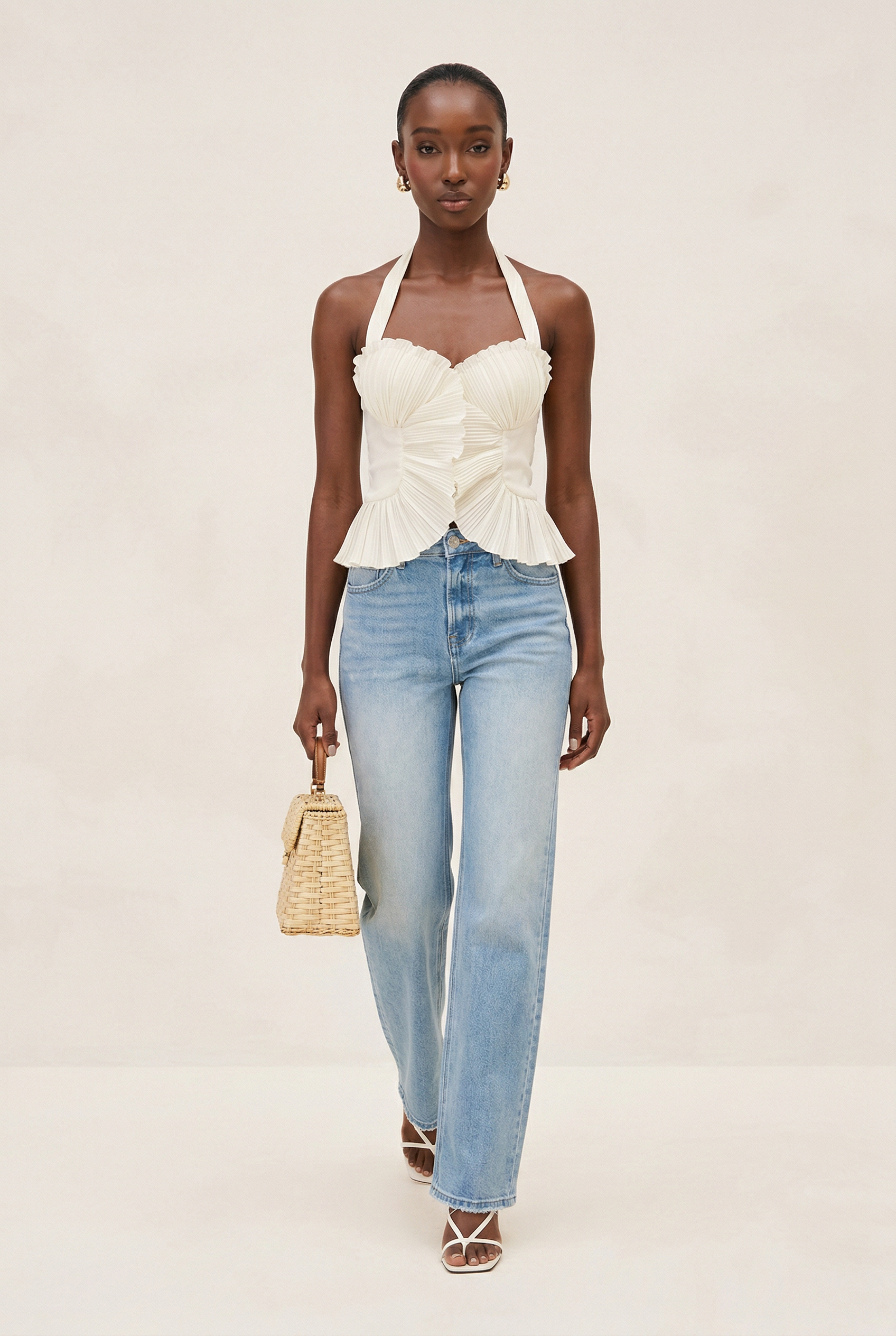 Woman wearing a white top and blue jeans with a woven handbag on a beige background