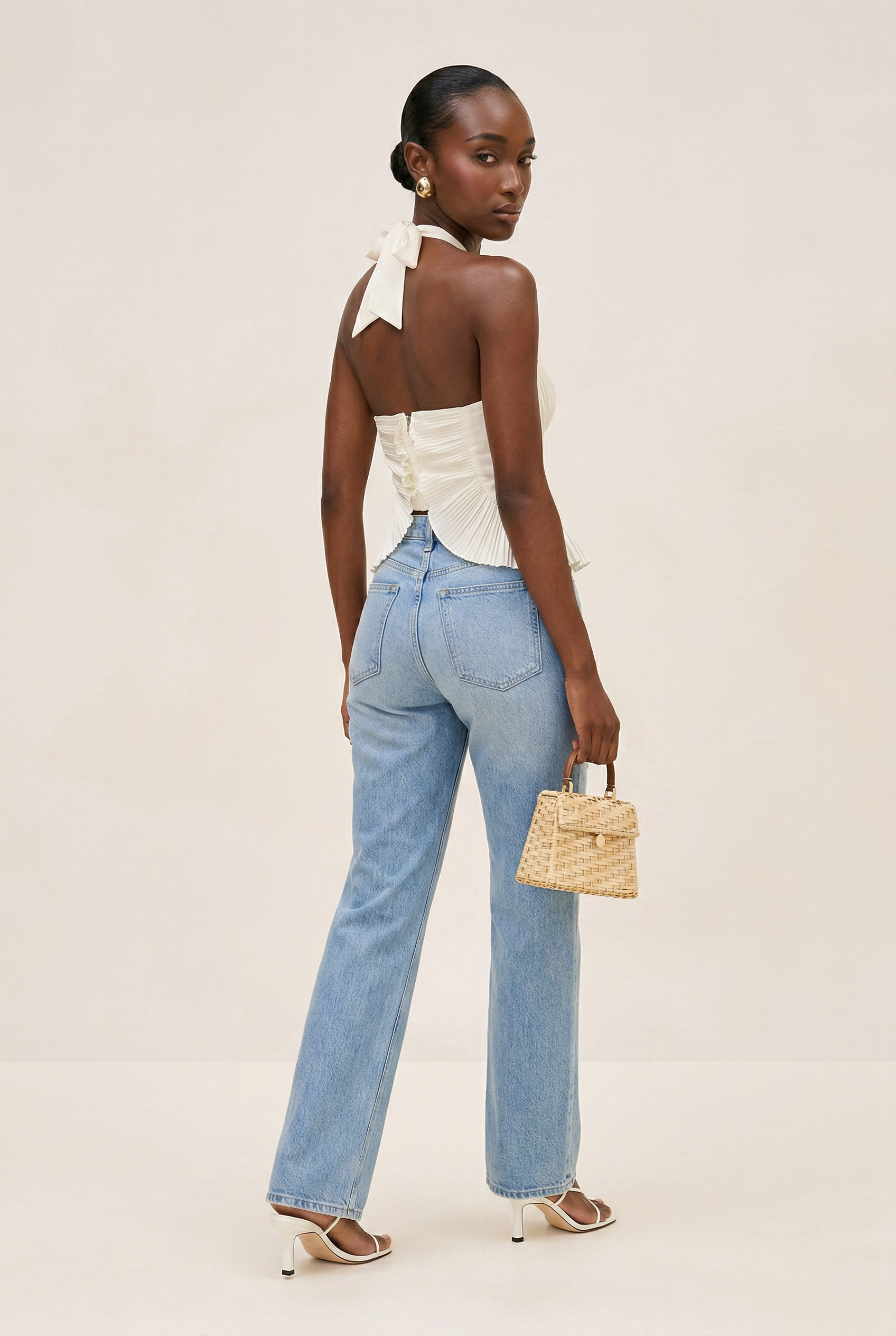 Woman wearing a white top and blue jeans, holding a woven handbag on a beige background