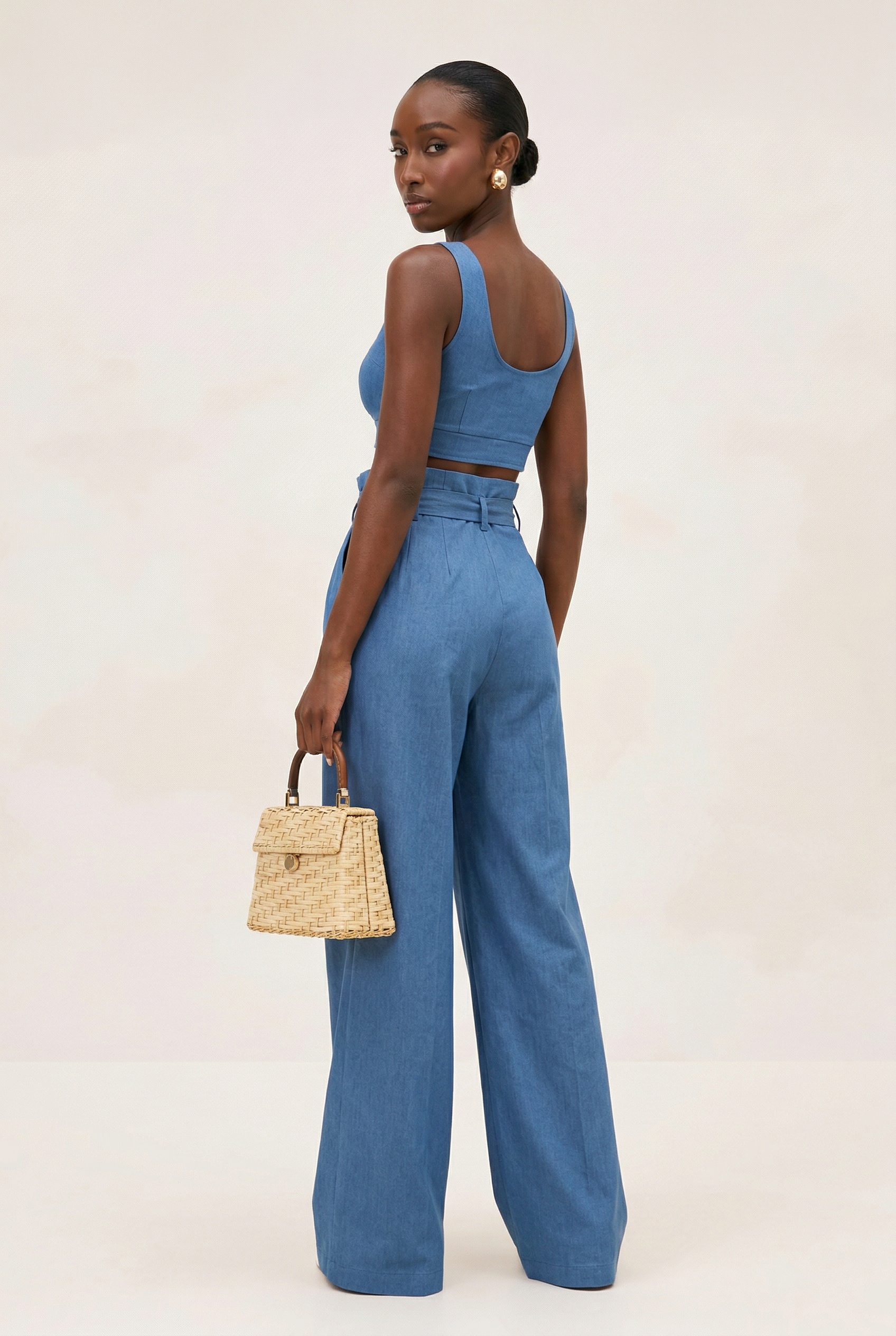 Woman wearing a blue jumpsuit holding a straw bag on a plain background