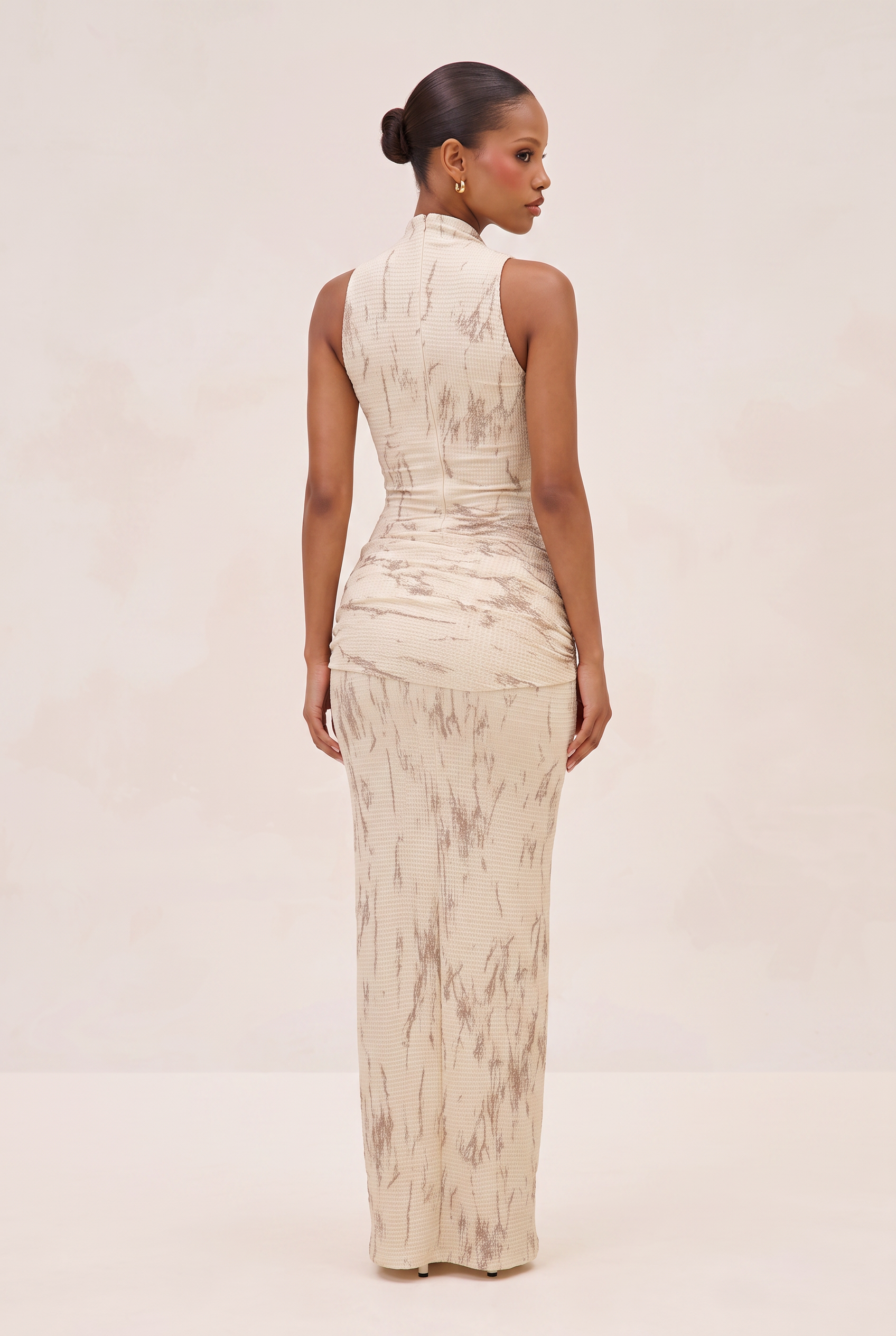 A woman shown from the back wearing a sleeveless, high-neck floor-length dress with a beige and taupe marbled pattern, set against a plain cream studio background.