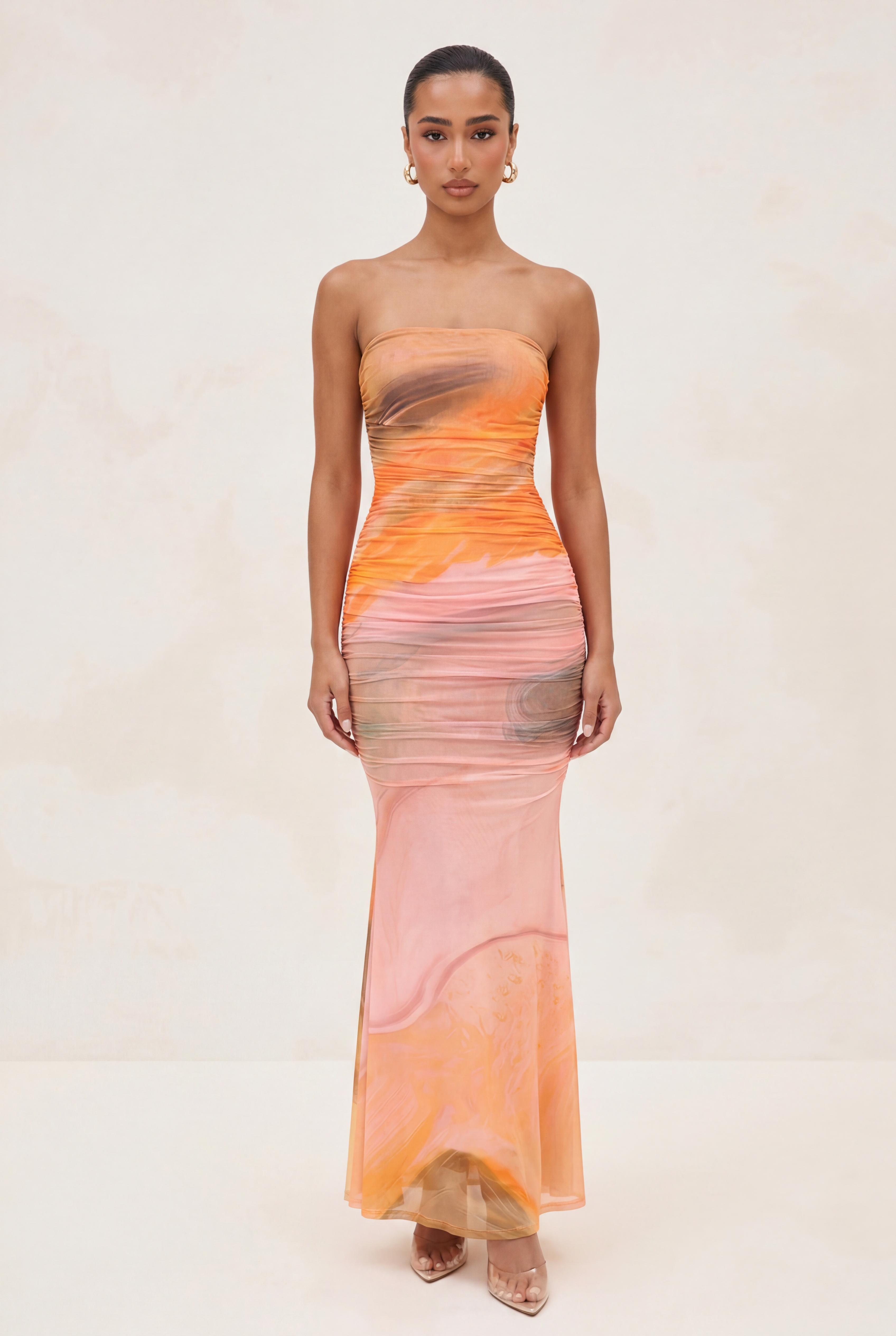 A woman modeling a strapless, floor-length mermaid gown with an abstract watercolor-like print in shades of orange, pink, peach, and beige against a light beige studio background.