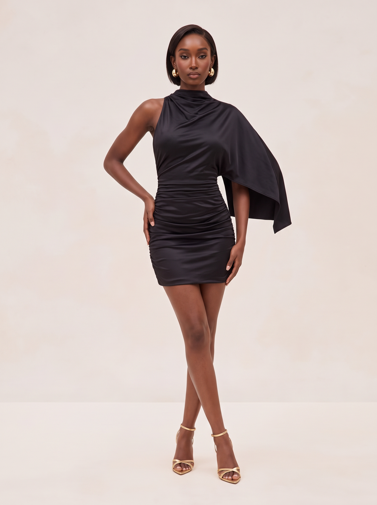 A woman modeling a black satin mini dress with a high draped neckline, one flowing cape-like sleeve, and gathered ruching at the waist, posed against a plain beige studio background.