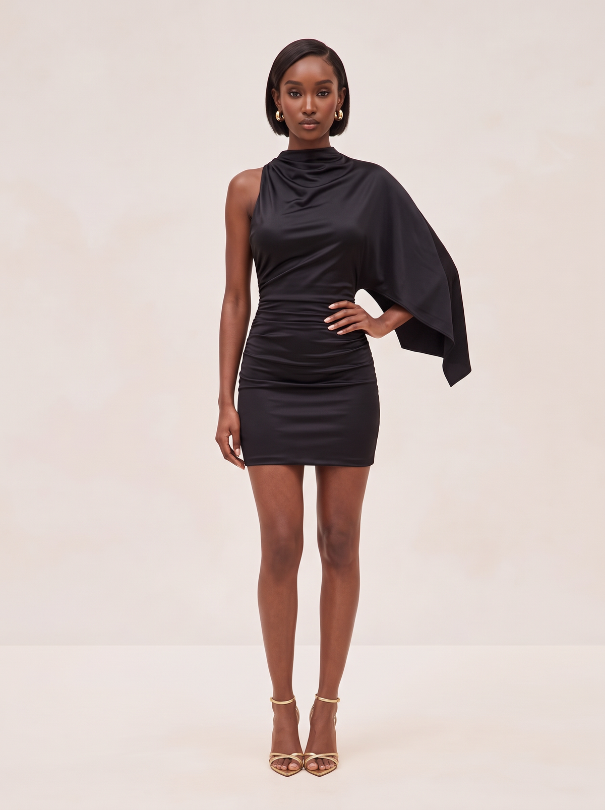A model wearing a black one-shoulder ruched mini dress with a draped cape sleeve, paired with gold high-heeled sandals, against a solid beige studio background.