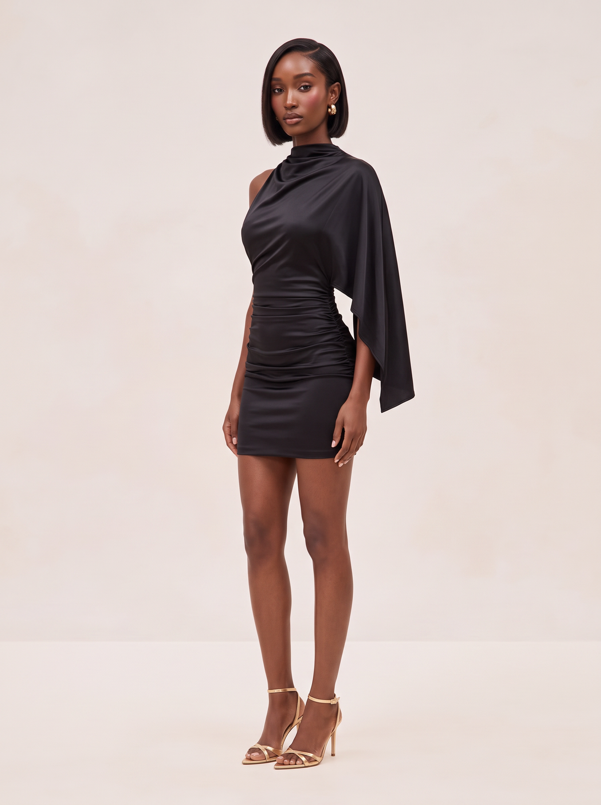A woman modeling a black satin mini dress with a draped capelet sleeve and ruched bodice, paired with gold strappy high-heeled sandals, against a solid light beige studio background.