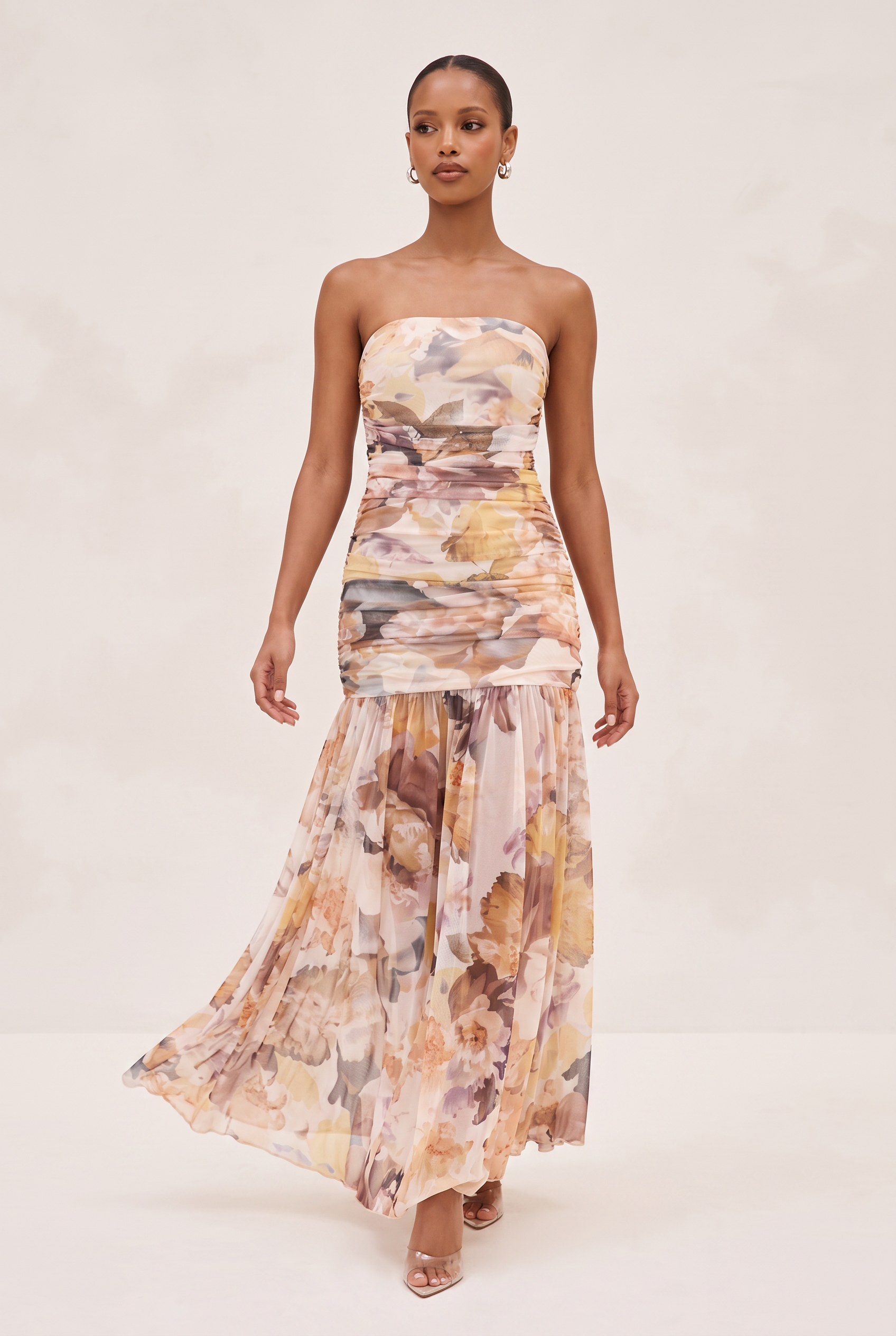 A woman modeling a strapless maxi dress with a ruched bodice and a flowing floral print skirt in soft beige, brown, and muted yellow tones, set against a plain light beige studio background.