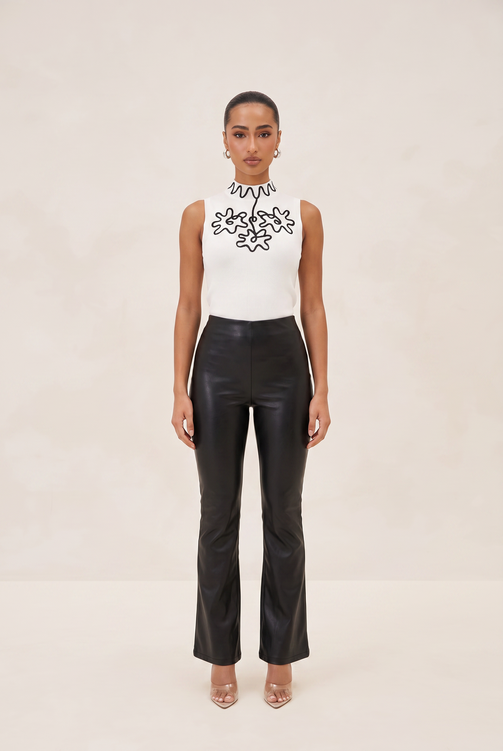Person wearing a white sleeveless top with black floral embroidery and black leather pants on a beige background