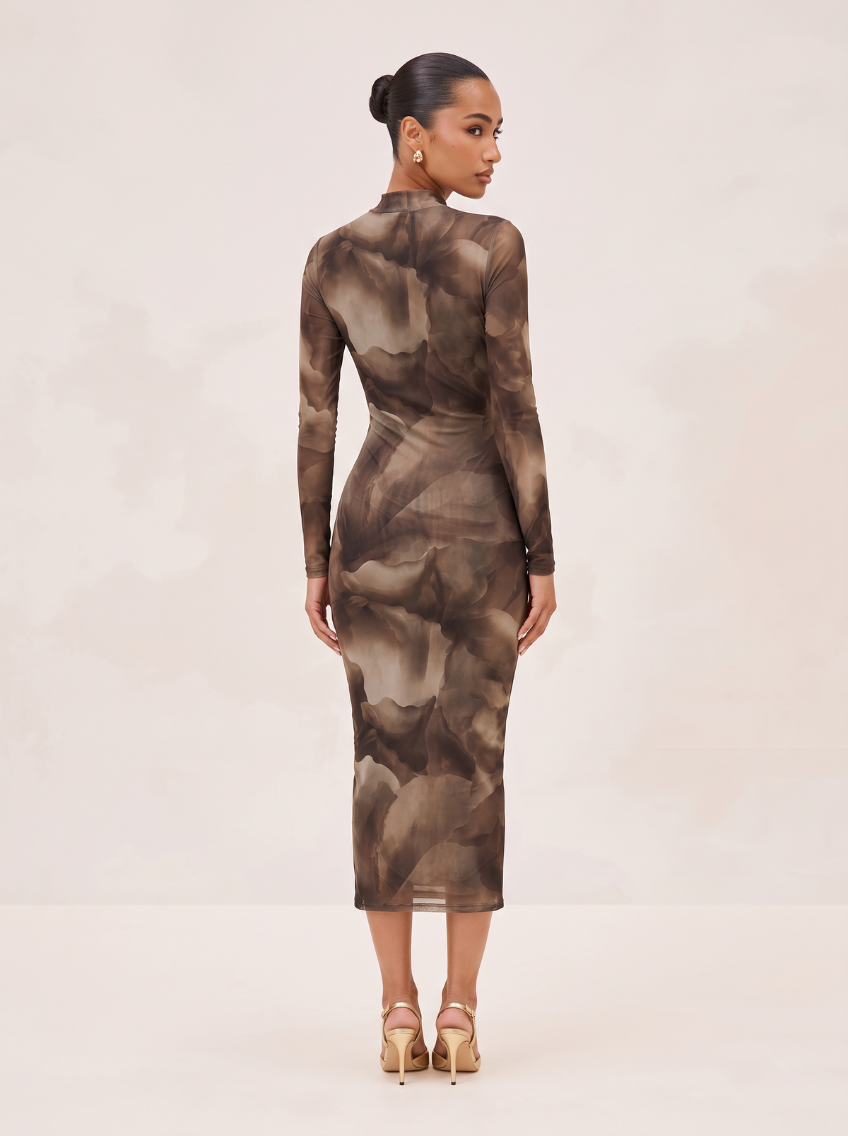 A woman stands in a studio wearing a fitted long-sleeve midi dress with an abstract brown and taupe watercolor-style print, shown from the back. She is wearing nude high-heeled sandals against a soft light beige background.