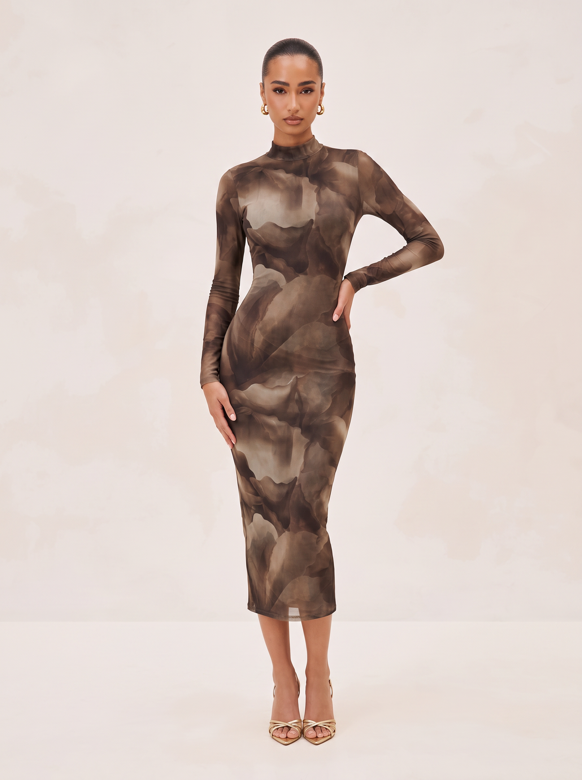 A woman modeling a long-sleeve mock-neck midi dress with an abstract watercolor-style print in shades of brown, taupe, and beige, paired with gold high-heeled sandals against a light beige studio background.