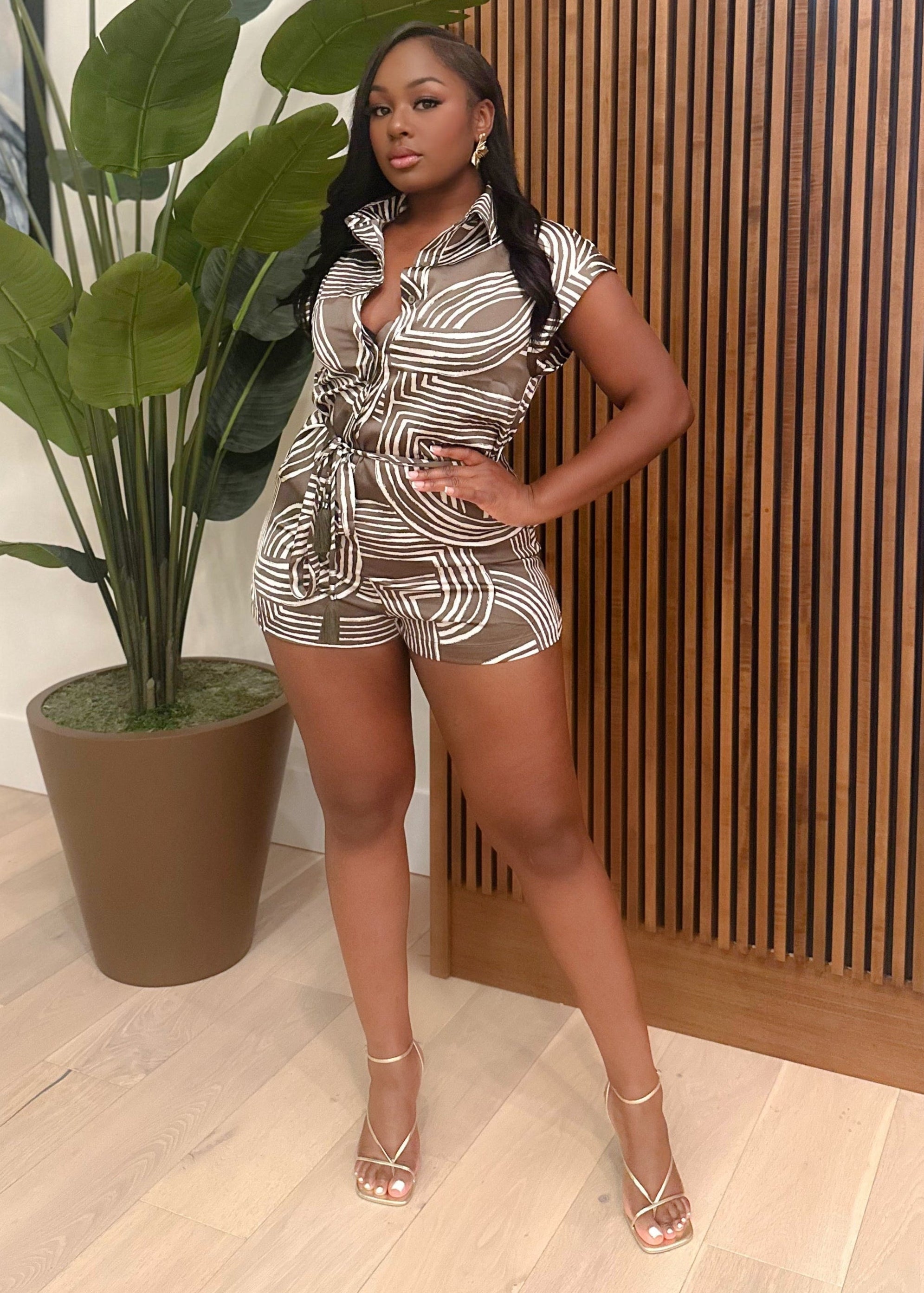 Woman in a patterned outfit standing indoors with a plant and wooden panel in the background