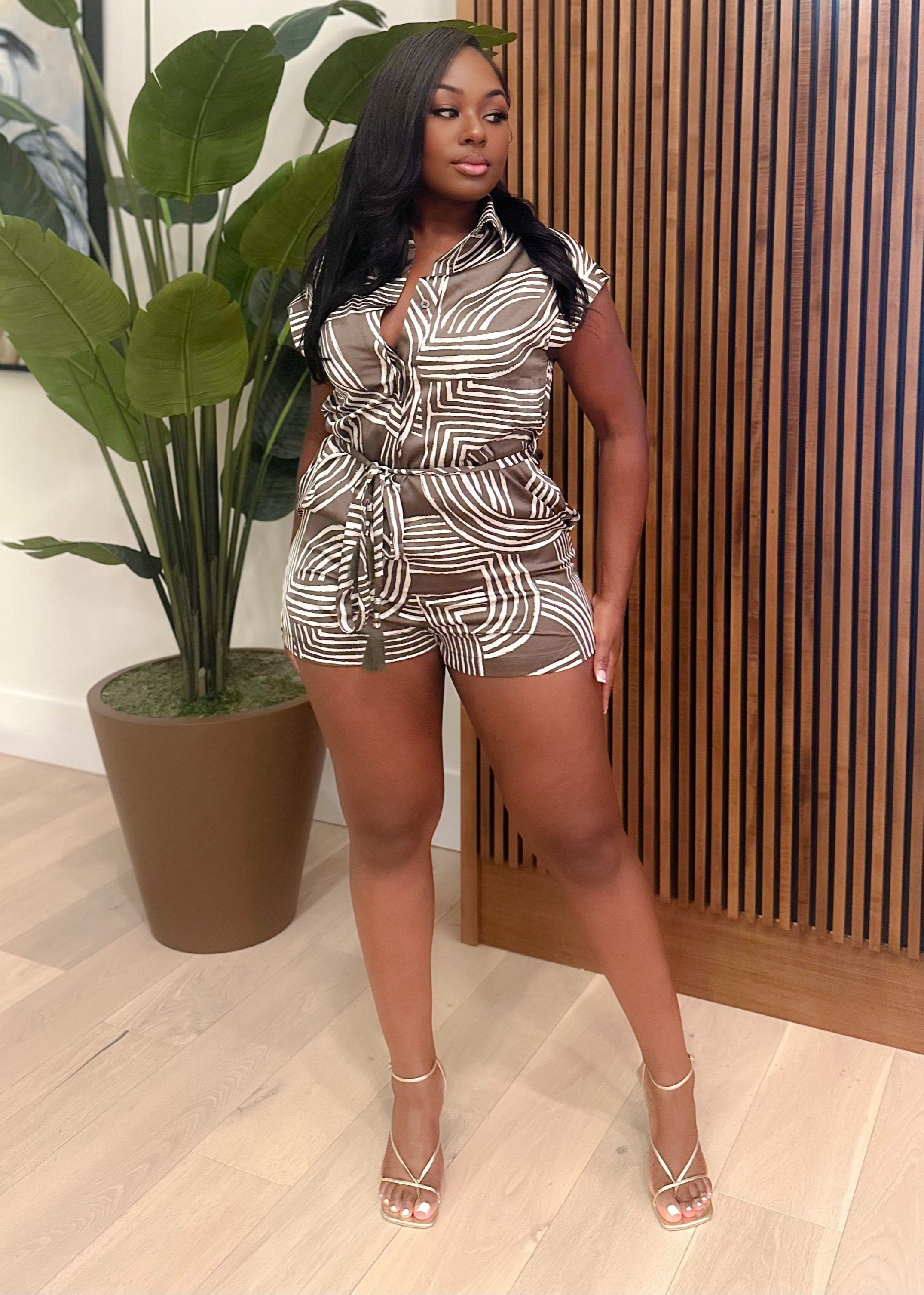 Woman in a patterned outfit standing indoors next to a plant and wooden wall.