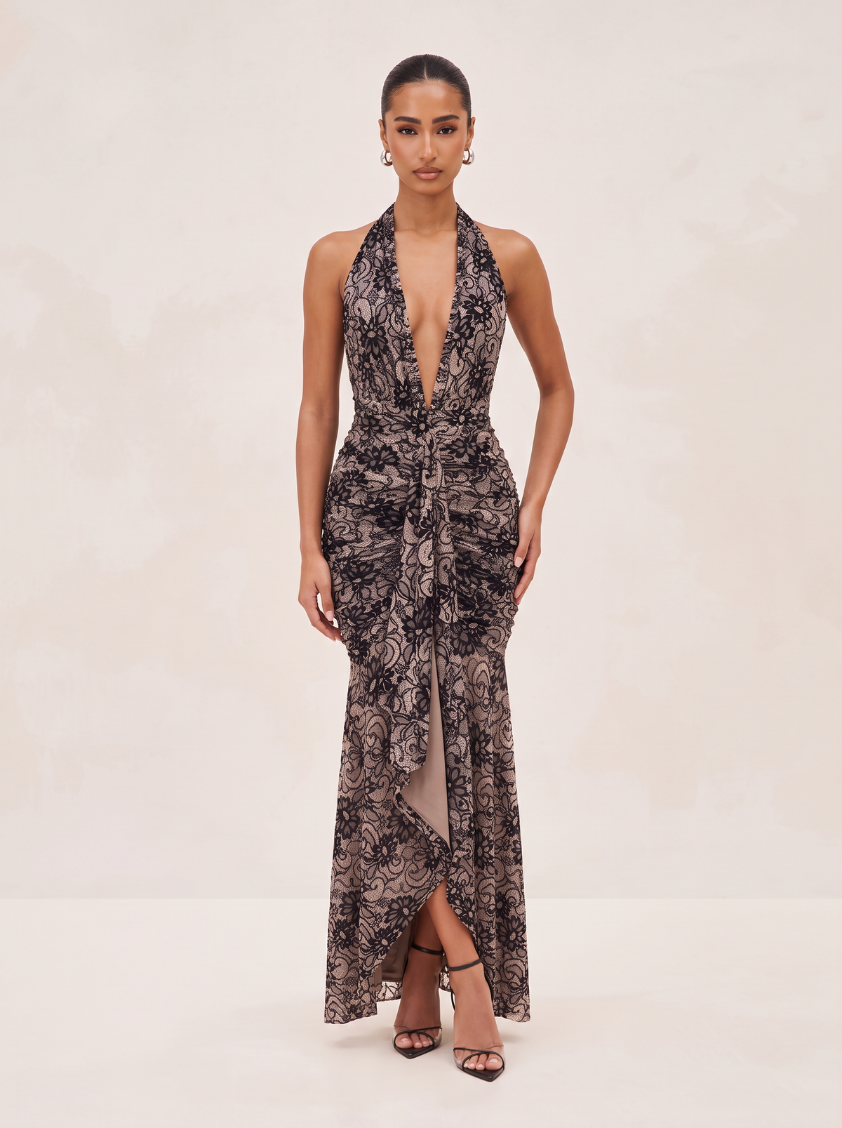 A model wearing a sleeveless halter-neck maxi dress with a deep plunging V-neckline, a wrap-style waist, and a sheer floral lace overlay. The dress features a high-low hem with a front slit, shown against a neutral cream studio background.
