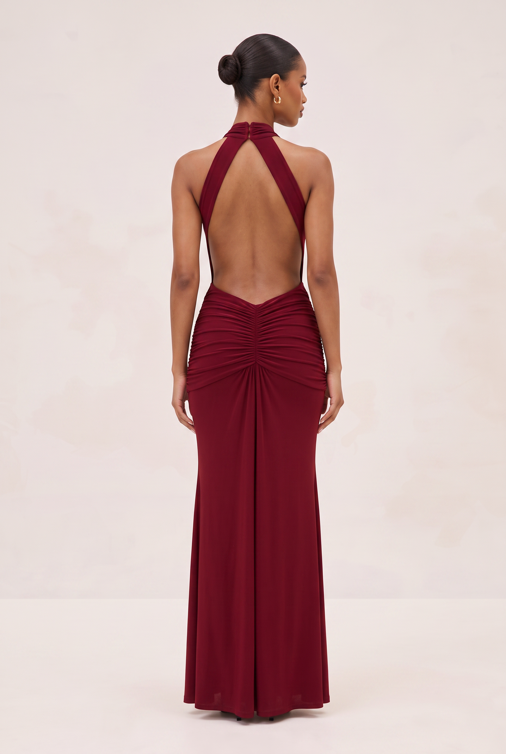 A woman shown from the back wearing a floor-length burgundy halter gown with an open back and gathered ruching at the waist, photographed against a plain light beige studio background.
