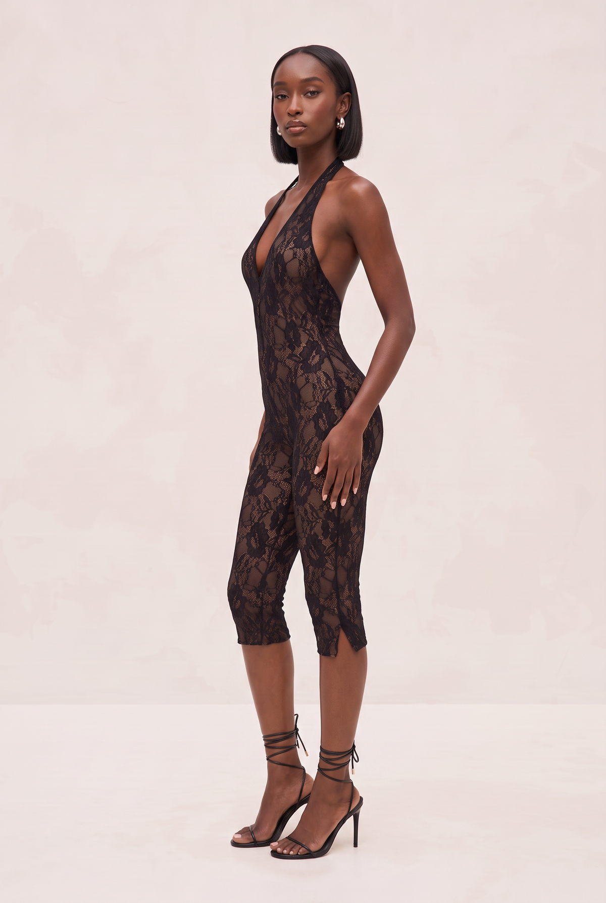 A woman modeling a black lace halter jumpsuit with a deep plunging neckline and capri-length legs, paired with black lace-up high heels, against a light beige studio backdrop.