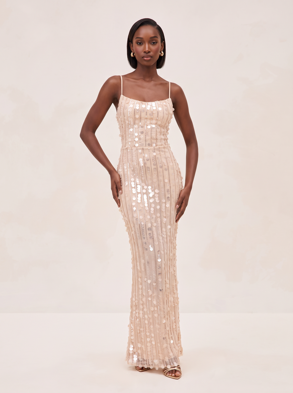 A woman modeling a floor-length champagne sequin gown with spaghetti straps and a fitted silhouette, standing against a soft beige studio backdrop.