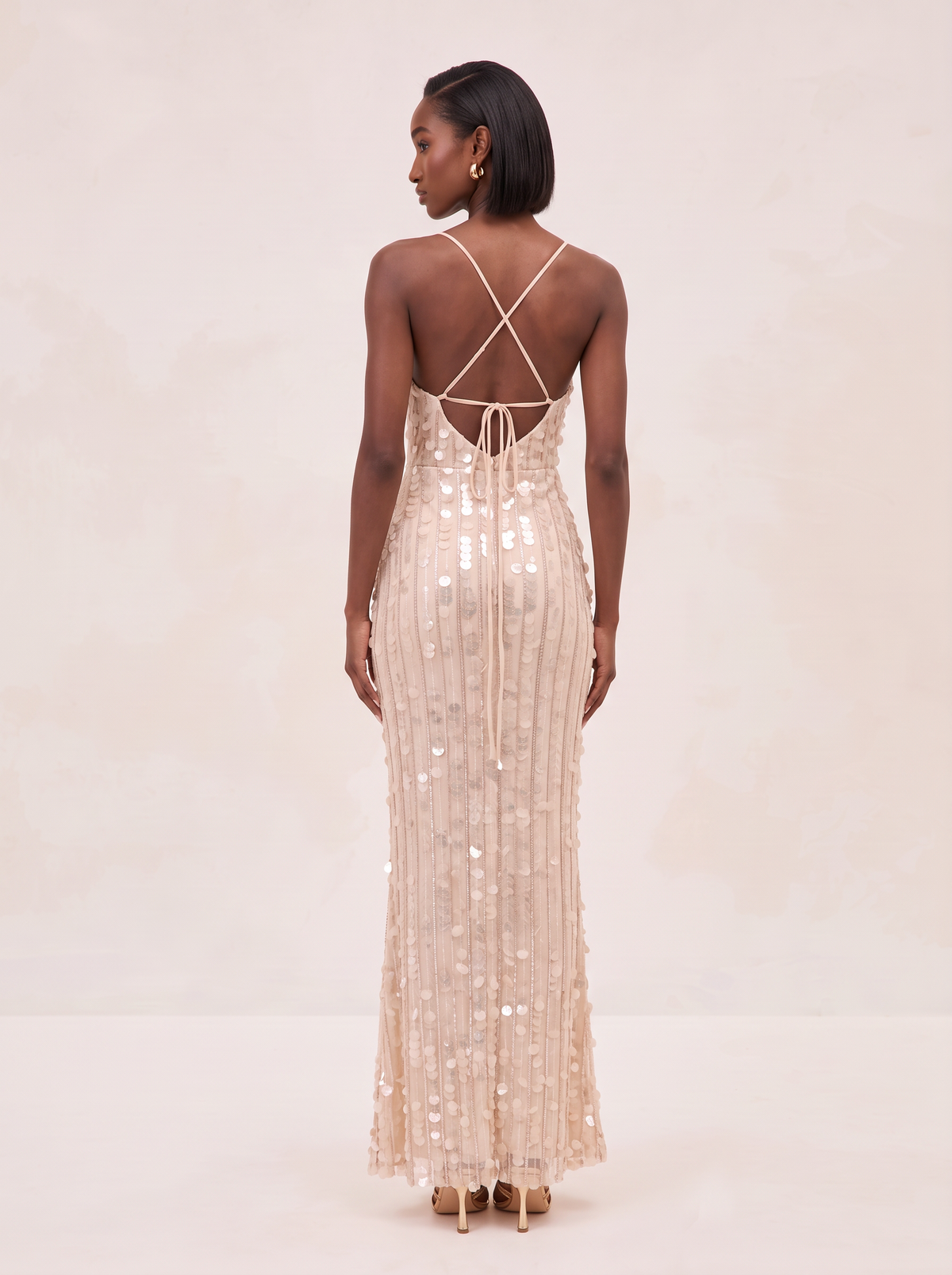 A woman wearing a floor-length champagne sequin gown, shown from the back against a warm beige studio background.