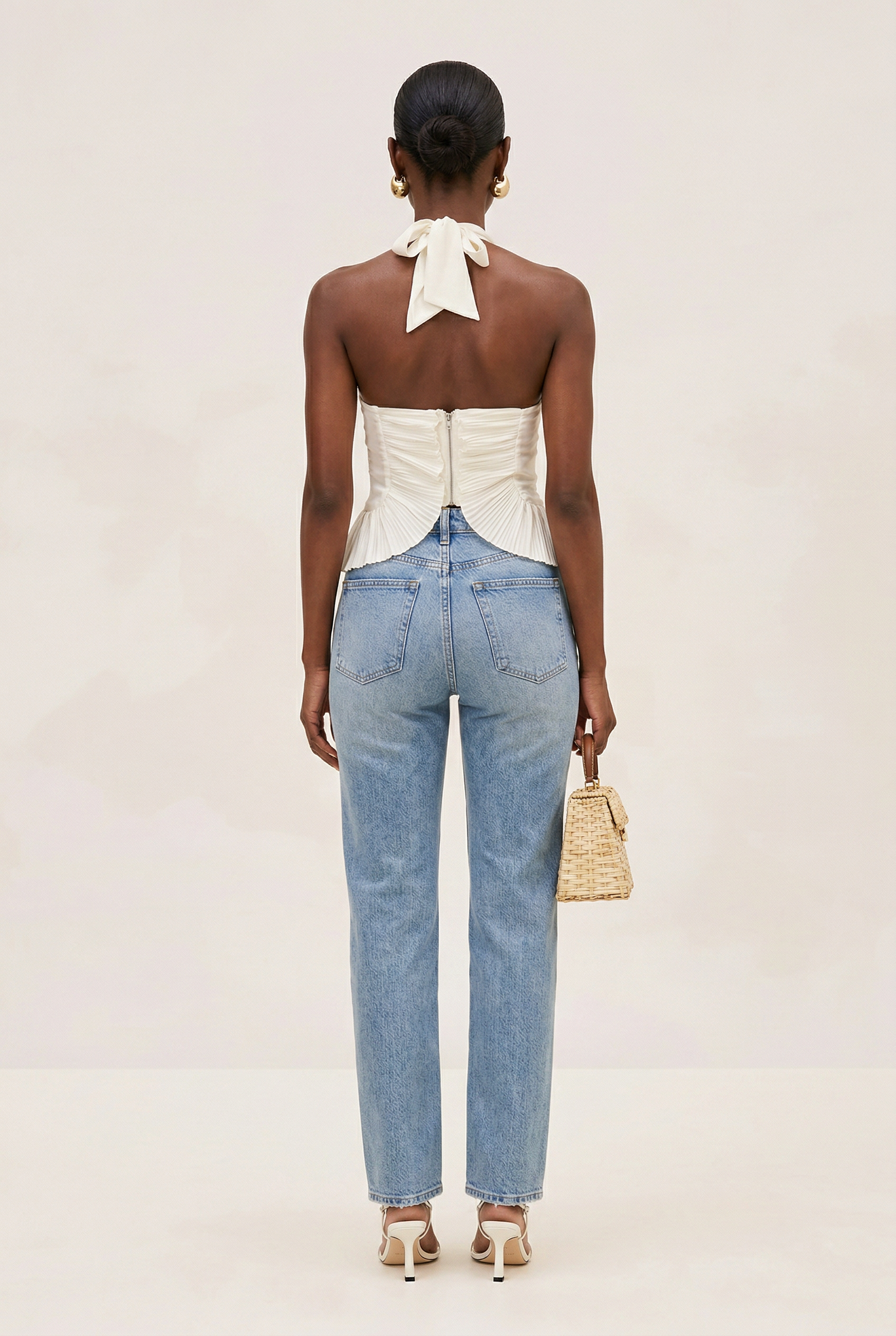 Woman wearing a white top with a bow detail and blue jeans, holding a straw bag on a plain background