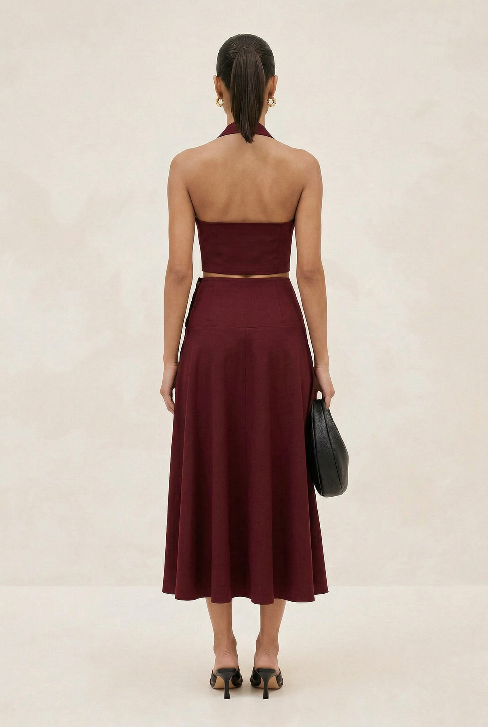 Woman wearing a burgundy strapless top and skirt set with a black clutch on a beige background