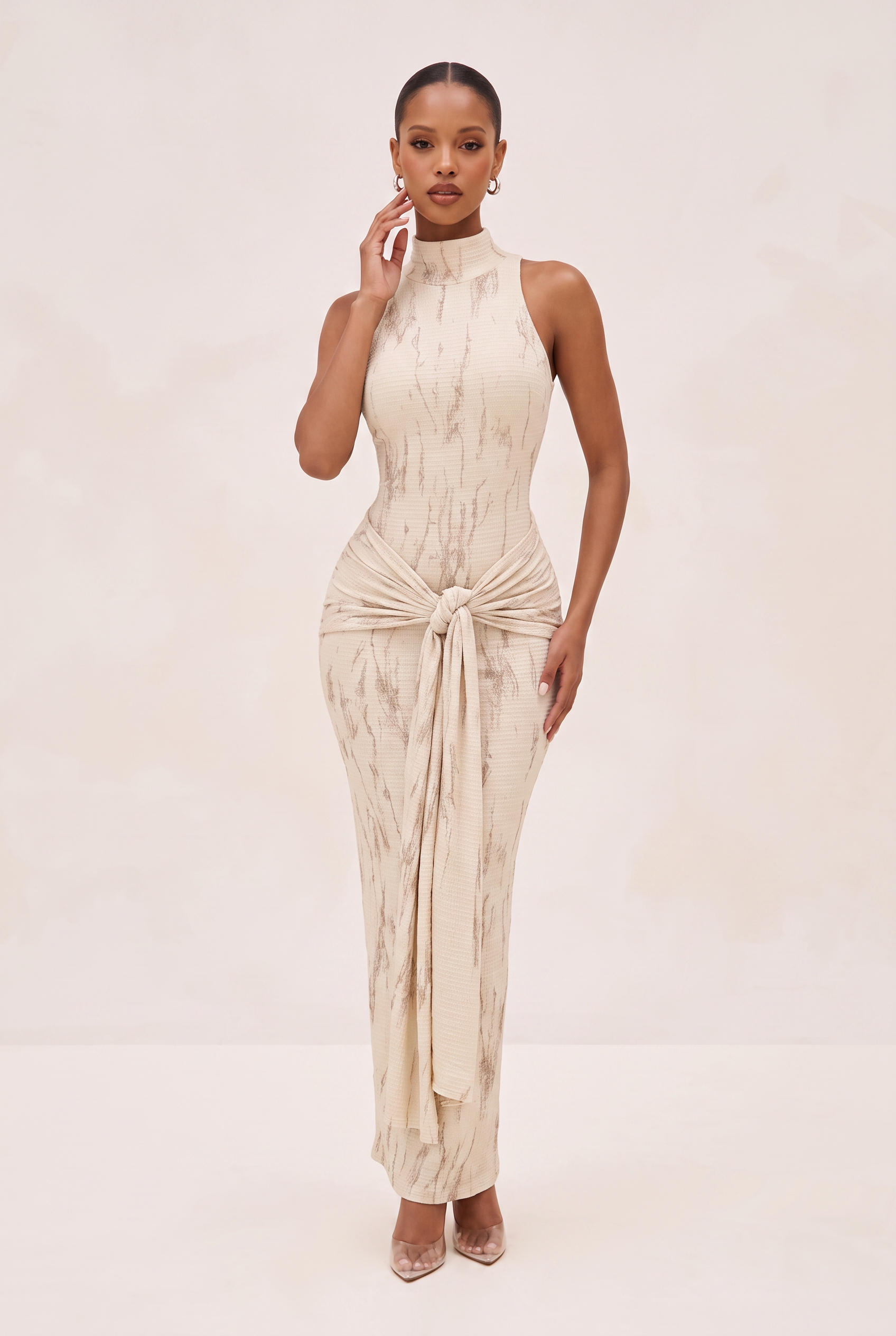 A woman modeling a sleeveless, high-neck, floor-length beige dress with a subtle marbled pattern and a gathered tie detail at the waist, set against a light beige studio background.