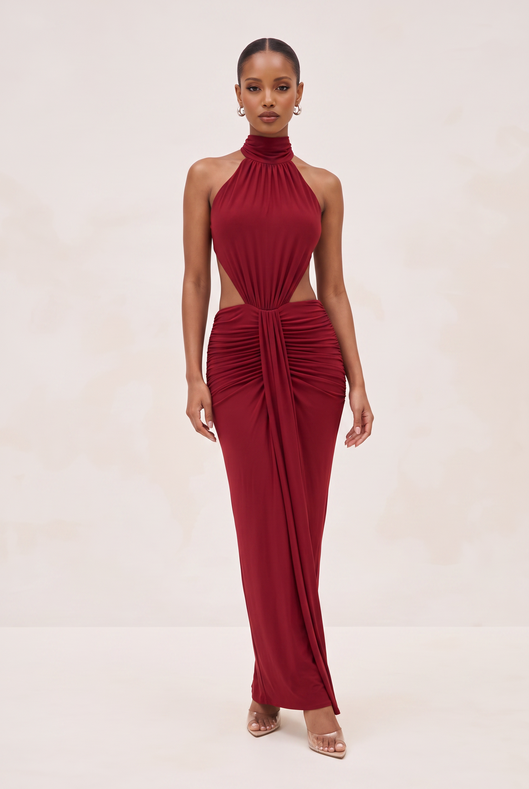 A woman modeling a sleeveless, high-neck, floor-length gown in deep burgundy red. The dress features a halter-style neckline, a side cutout at the waist, and dramatic gathered draping around the waist and cascading down the skirt.