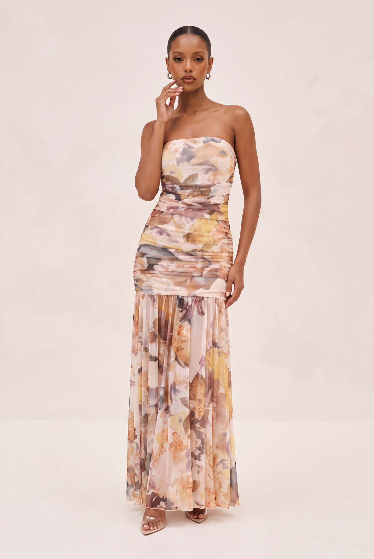 A woman modeling a strapless maxi dress with a floral watercolor print in soft beige, brown, and muted orange tones, standing against a plain light beige studio background.