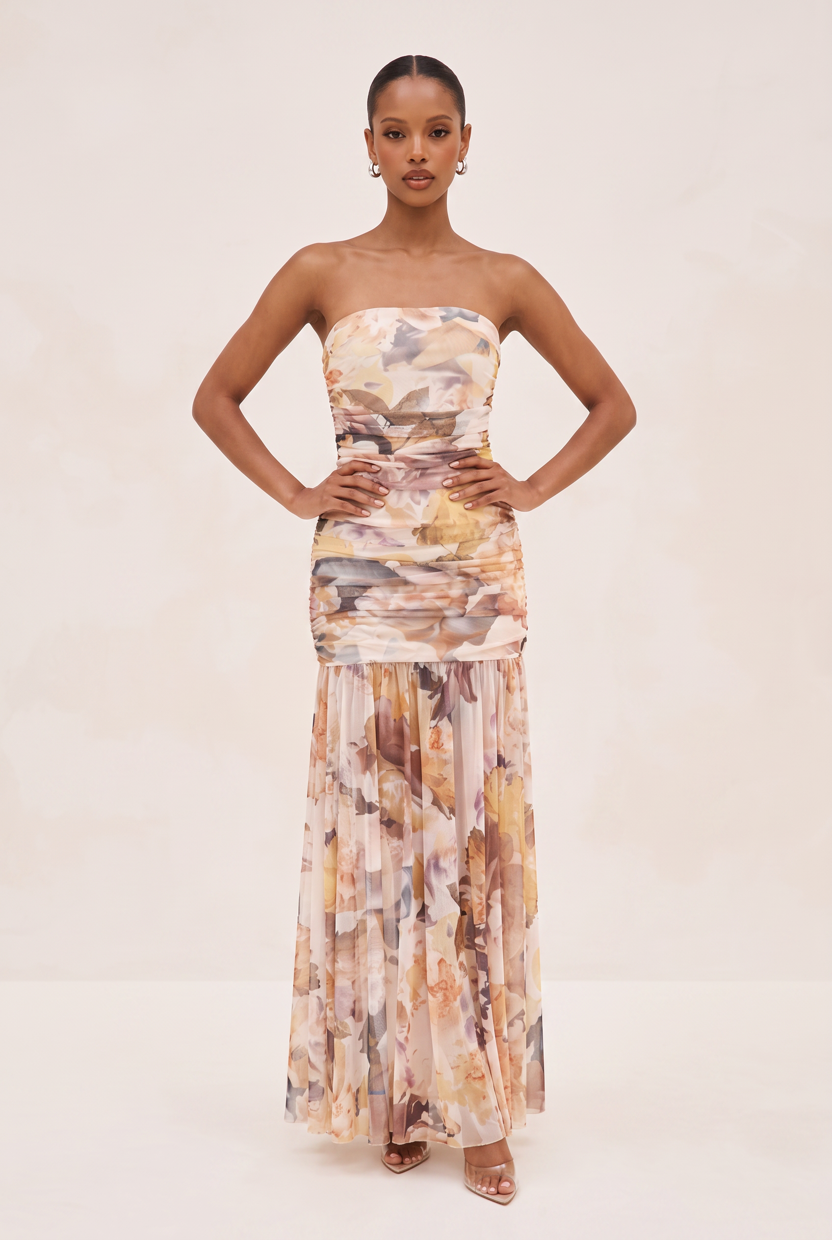 A woman modeling a strapless maxi dress with an abstract painterly print in beige, brown, and muted orange tones, set against a plain cream studio background.