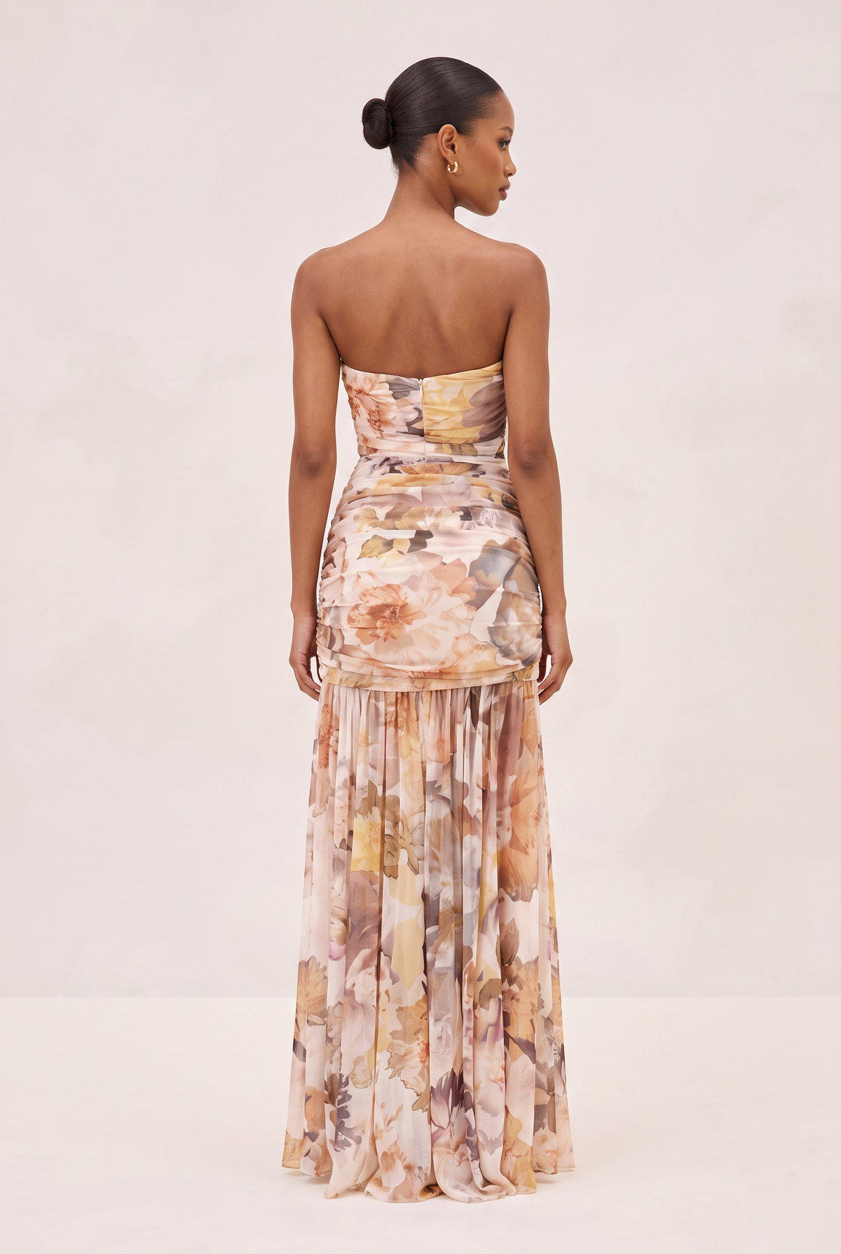 A woman stands with her back to the camera wearing a strapless floor-length gown with a soft abstract floral print in blush, beige, tan, and muted brown tones against a plain light studio background.