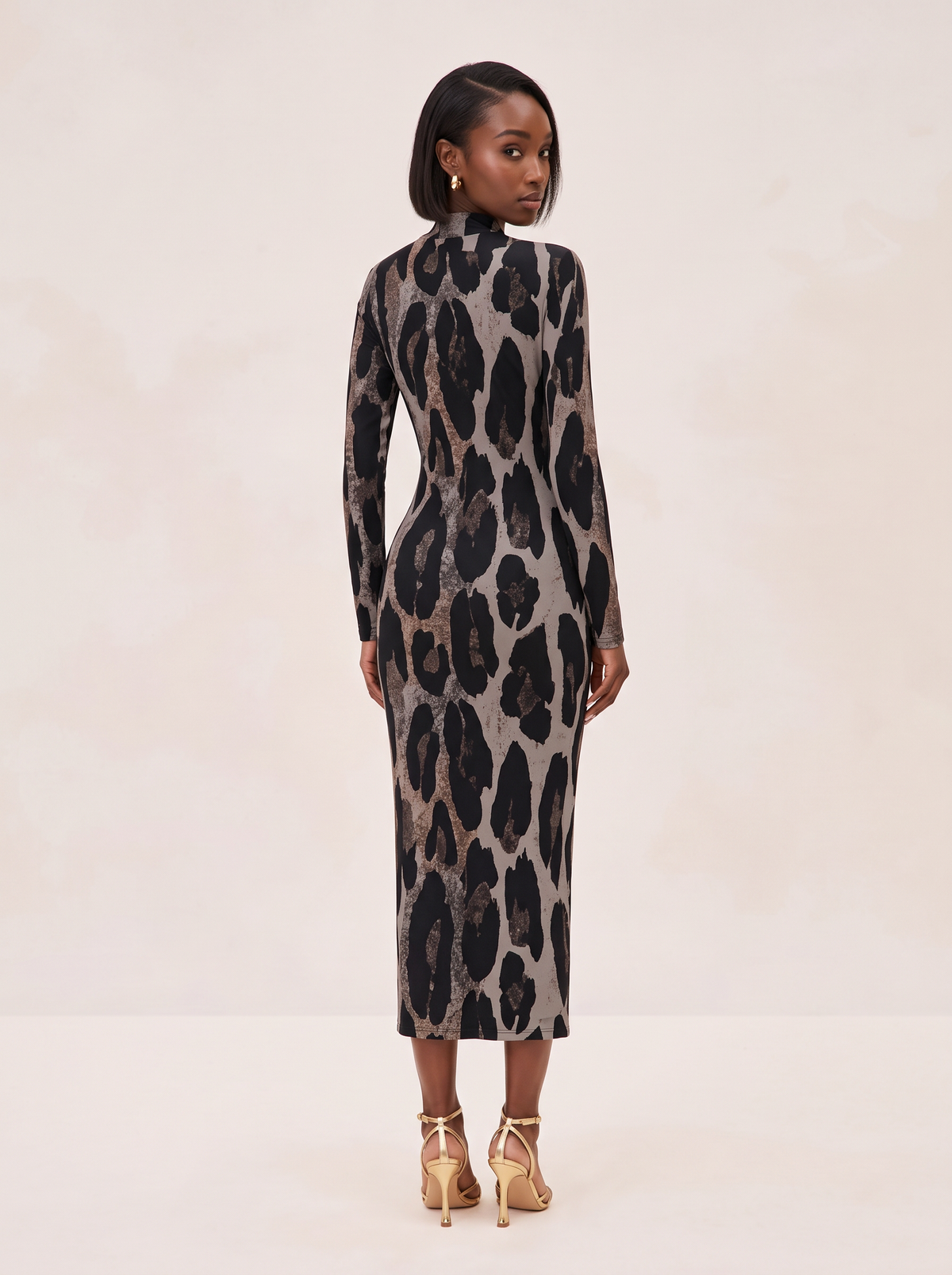 A woman stands with her back to the camera wearing a long-sleeve mock-neck midi dress with a black and brown leopard print on a beige background, paired with gold high-heeled sandals, against a light beige studio backdrop.