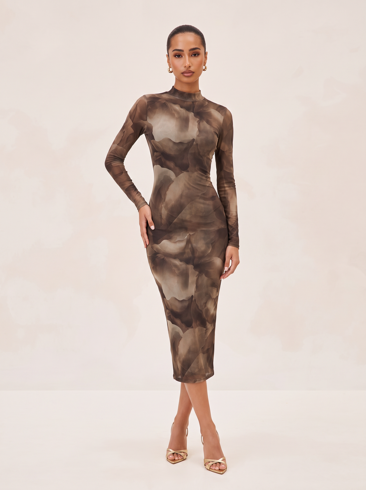 A woman modeling a long-sleeve mock-neck midi dress with an abstract brown and beige floral pattern, standing against a light beige studio background.
