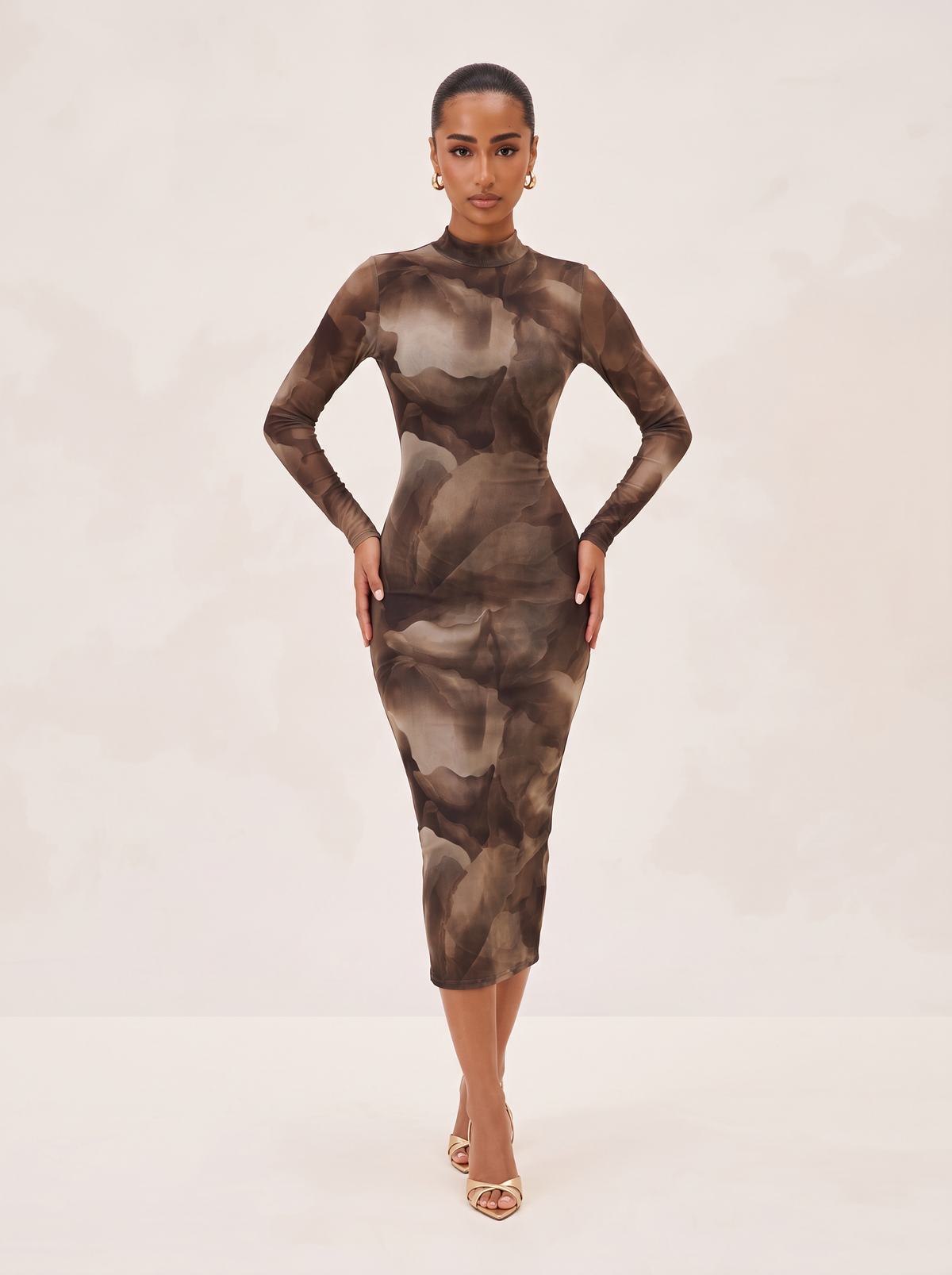 A model wearing a long-sleeve mock-neck midi dress with an abstract watercolor pattern in shades of brown, beige, and gray, paired with gold strappy heels against a light beige studio background.