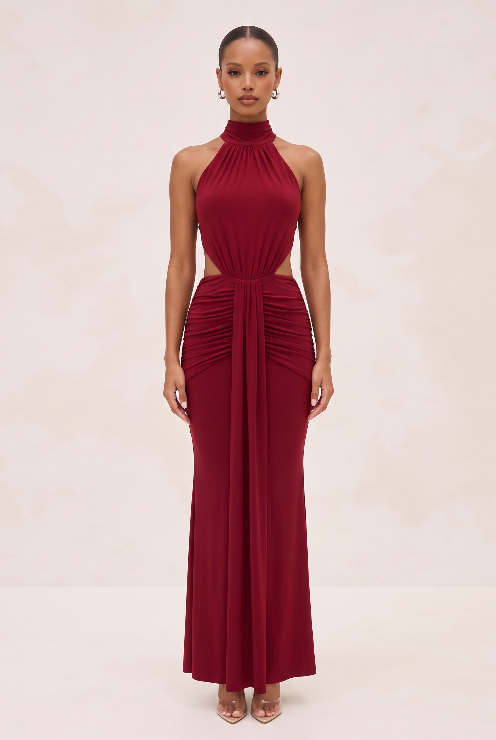 A woman modeling a sleeveless, high-neck, floor-length gown in deep burgundy. The dress features a structured bodice, side cutouts, and a gathered, ruched skirt.
